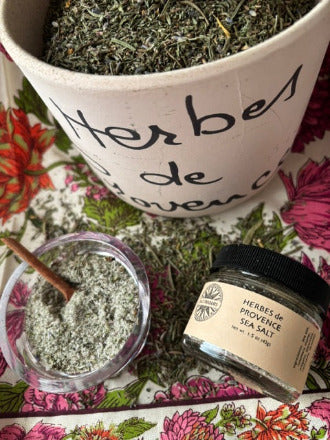 A styled flat-lay of a herbal product set featuring a large earthenware pot labeled 'Herbes de Provence' filled with dried green herbs, a small glass jar of 'Herbes de Provence Sea Salt' with a black lid, and a small glass bowl of the sea salt with a wooden spoon, all arranged on a pink and green floral patterned fabric.