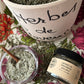 A styled flat-lay of a herbal product set featuring a large earthenware pot labeled 'Herbes de Provence' filled with dried green herbs, a small glass jar of 'Herbes de Provence Sea Salt' with a black lid, and a small glass bowl of the sea salt with a wooden spoon, all arranged on a pink and green floral patterned fabric.